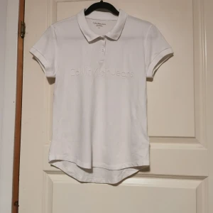 Vit pikétröja från Calvin Klein Jeans - Säljer en stilren vit pikétröja från Calvin Klein Jeans i storlek S. Tröjan har korta ärmar och en klassisk krage med knappar. Perfekt för en avslappnad men ändå snygg look. Passar både till vardags och lite finare tillfällen.