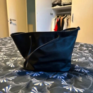 Svart axelväska med textmönster - Säljer en stilren svart axelväska i syntetmaterial. Väskan har ett coolt textmönster på insidan med namn på olika städer som Milano och Berlin. Perfekt för vardagsbruk eller en dag på stan. Den är rymlig och har långa handtag för enkel bärning. Passar till alla outfits!