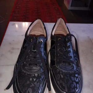 Svarta sneakers till dam - Snygga svarta sneakers med dekorativt leopardmönster. De har snörning och en bekväm passform, perfekta för både vardag och fest. Skorna är i mycket bra skick och har en stilren design som passar till det mesta.