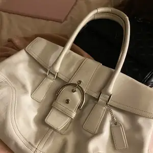 Säljer en elegant vit handväska från Coach i skinn. Väskan har en stilren design med ett dekorativt bälte och en praktisk dragkedja inuti. Perfekt för både vardag och fest! 👜✨