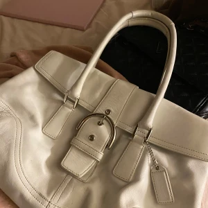 Vit handväska från Coach - Säljer en elegant vit handväska från Coach i skinn. Väskan har en stilren design med ett dekorativt bälte och en praktisk dragkedja inuti. Perfekt för både vardag och fest! 👜✨
