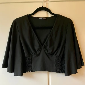 Svart croppad topp från Shein - Säljer en svart croppad topp från Shein i storlek M. Toppen har en v-ringad design och vida ärmar som ger en snygg look. Den har en smockad detalj vid midjan för en bekväm passform. Perfekt för både vardag och fest!