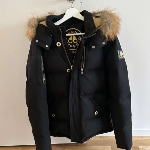 Mooseknuckles jacka NYskick* - Säljer en svart dunjacka från Moose Knuckles i mycket bra skick. Jackan har en fluffig pälsdetalj på huvan och är perfekt för kalla vinterdagar. Den har dragkedja och knappar framtill samt långa ärmar. Perfekt för att hålla sig varm och stilren! ”Unik med guld detaljer ” nypris 14000