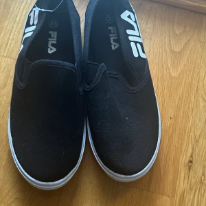 Svarta slip-on sneakers från Fila - Säljer ett par svarta slip-on sneakers från Fila i mycket bra skick. De har en enkel och stilren design med vit sula och Fila-loggan på insidan. Perfekta för vardagsbruk och lätta att matcha med olika outfits.