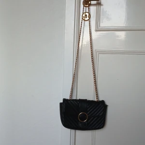 Svart axelväska med guldkedja - Snygg svart axelväska med en guldkedja. Väskan har en stor guldring som detalj på framsidan. Den har en lång kedja sp att man både kan ha den över kroppen, men man kan också vika kedjan dubbel och ha med ett kortare band! Den stängs med en magnetisk "knapp" och har ett fack inuti med dragkedja. Som visat på bilderna har färgen försvunnit lite på kanterna av väskan, men det är inget som syns lång bort!🥰