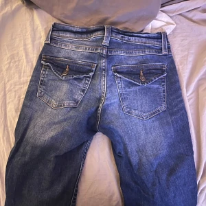 Blå jeans med knappar och fickor  - Säljer ett par snygga blå jeans i bra skick. De har en klassisk femficksdesign med knappar på bakfickorna. Jeansen har en normal passform och är perfekta för vardagsbruk. Passar till både sneakers och boots!