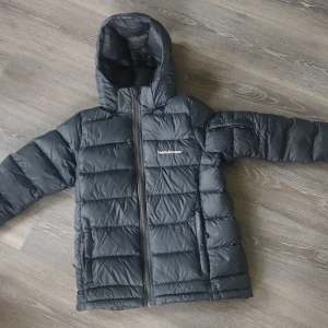 Svart dunjacka från Peak Performance, 160 - Säljer en svart dunjacka från Peak Performance i bra skick. Jackan har en praktisk dragkedja och är perfekt för kalla vinterdagar. Den är långärmad och har en skön huva för extra värme. Perfekt för både vardag och utomhusaktiviteter! Finns en liten reva på ena armbågen och en av fickornas dragkedja fungerar inte.