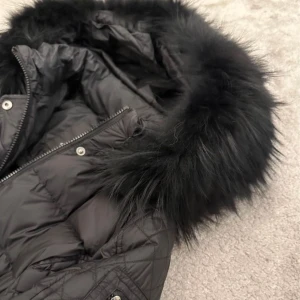 Svart dunjacka från Rockandblue - Säljer en snygg svart dunjacka från Rockandblue i storlek 40. Jackan har en fluffig pälsdetalj på huvan och quiltade mönster på sidorna. Den är perfekt för kalla vinterdagar och har både dragkedja och knappar för extra värme. Superbra skick och redo för nya äventyr! 🖤 modell Mid Beam