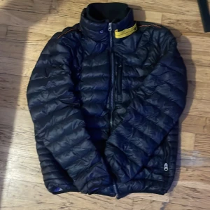 Svart pufferjacka från Parajumpers - Säljer en svart pufferjacka från Parajumpers i superlätt material. Jackan har en dragkedja framtill och en hög krage med en gul detalj. Perfekt för kyliga dagar och håller dig varm utan att kännas tung. Passar både till vardags och lite mer äventyrliga aktiviteter. Har använt den några gånger bara, inga skador på jackan.