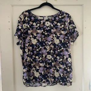 Blommig topp från ICHI - Säljer en superfin blommig topp från ICHI i storlek 40. Den har en mörk bas med blommor i lila, beige och vit. Toppen är kortärmad och perfekt för både vardag och fest. Materialet känns lätt och luftigt, vilket gör den skön att bära.