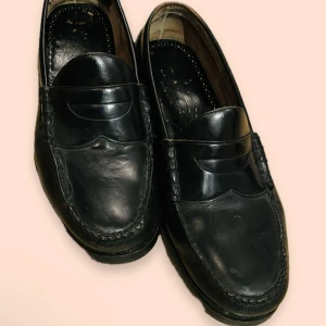 Svarta loafers läder - Säljer ett par klassiska svarta loafers i läder. De har en stilren design med en dekorativ rem över vristen. Skorna är i välanvänt skick men välbehandlade och bekväma. Skorna har tagits till skomakare nyligen för polering och ky klack och sula!!