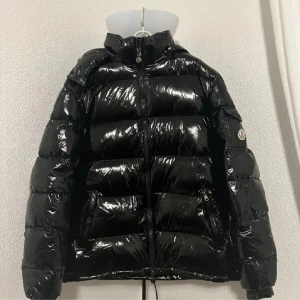 Svart glansig pufferjacka från Moncler - Säljer en supercool Moncler Maya. Den är glansig och har en dragkedja framtill. Jackan är perfekt för vintern och håller dig varm och stilren. Den har långa ärmar och en snygg logga på ärmen. Den är storlek 5 som är xl/L