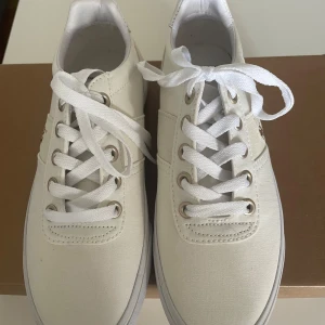 Vita sneakers från Polo Ralph Lauren - Säljer ett par snygga vita sneakers från Polo Ralph Lauren. De har en klassisk design med snörning och är gjorda i ett stilrent syntetmaterial. Perfekta för både vardag och fest!
