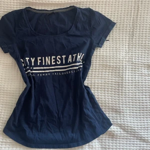 Mörkblå t-shirt från Gina Tricot - Säljer en snygg mörkblå t-shirt från Gina Tricot i storlek XS. Den har ett tryck på framsidan med texten 'CITY FINEST ATHL'. Perfekt för en avslappnad stil. T-shirten är kortärmad och gjord i ett mjukt material.