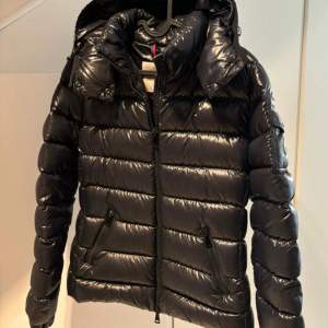 Säljer en snygg svart pufferjacka från Moncler. Den är perfekt för vintern med sitt quiltade mönster och långa ärmar. Jackan har en dragkedja framtill och en huva för extra värme. Moncler-loggan syns tydligt på ärmen. Den är i nyskick och redo för kalla dagar!