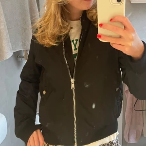 Svart bomberjacka - Säljer en stilren svart bomberjacka med dragkedja framtill. Jackan har en klassisk design med långa ärmar och ribbade muddar vid ärmslut och nederkant. Perfekt för en cool och avslappnad look.
