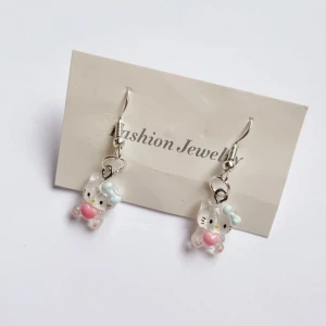 Söta örhängen med kattmotiv💓 - Charmiga örhängen med genomskinliga kattfigurer som har blå rosetter och rosa hjärtan. Perfekta för att lägga till en lekfull touch till din outfit. De hänger från silverfärgade krokar och är lätta att bära.🌸