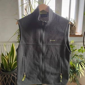 Grå fleeceväst från Patagonia - Säljer en grå fleeceväst från Patagonia med dragkedja framtill och två praktiska fickor. Perfekt för lager-på-lager under kyligare dagar. Västen har en enkel och stilren design.