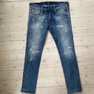 Blå jeansbyxor från Replay - Snygga blå jeansbyxor från Replay med slitna detaljer och en klassisk femficksdesign. Byxorna har en straight passform och är perfekta för en avslappnad stil. De har en knappgylf och en läderpatch med Replay-logga på baksidan.
