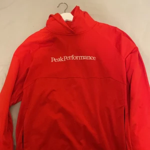 Röd hoodie från Peak Performance - Säljer en snygg röd hoodie från Peak Performance. Den har en klassisk design med logotypen tryckt på bröstet och en praktisk ficka framtill. Perfekt för en avslappnad stil.