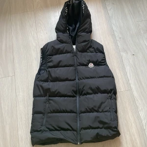 Svart dunväst från Moncler - Säljer en snygg svart dunväst från Moncler med huva och dragkedja. Västen har ett stilrent moncler märke på bröstet och praktiska fickor med dragkedja. Perfekt för kyligare dagar när du vill hålla stilen. 🖤(kan sänka pris rejält om det blir snabb affär)