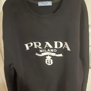 Svart tröja från Prada - Snygg svart tröja från Prada med deras ikoniska logga tryckt på framsidan. Tröjan har en klassisk rund hals och långa ärmar, perfekt för en stilren look.