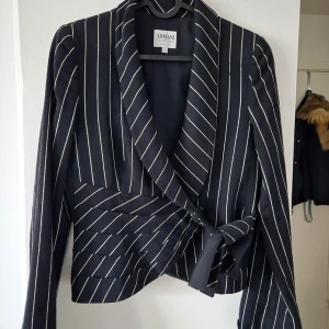 Svart randig kavaj från Armani Collezioni - Snygg svart kavaj från Armani Collezioni med vita ränder och en elegant knytning framtill. Kavajen har en klassisk design med långa ärmar och en figursydd passform. Perfekt för en stilren look.