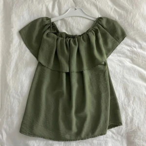 Olivgrön offshoulder topp - Säljer en stilren olivgrön offshoulder topp med en luftig passform. Perfekt för varma dagar och kan enkelt matchas med olika outfits. Toppen har en bred volangdetalj runt axlarna som ger en extra touch av elegans.