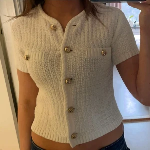 Vit stickad kortärmad topp med knappar - Säljer en stilren vit stickad topp med korta ärmar och dekorativa guldknappar framtill. Toppen har en ribbad struktur och två små fickor på bröstet. Perfekt för en chic och avslappnad look.