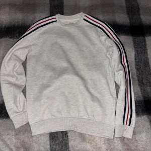 Grå sweatshirt från YOURTURN - Säljer en stilren grå sweatshirt från YOURTURN med randiga detaljer i rött, vitt och svart längs ärmarna. Tröjan har en klassisk passform och är perfekt för en avslappnad look. Den är tillverkad i ett mjukt material som ger en skön känsla.