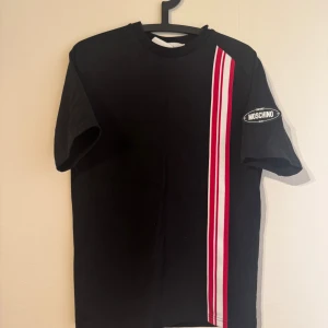 Svart t-shirt från Moschino med ränder - Snygg svart t-shirt från Moschino med röda och vita ränder på framsidan. T-shirten har korta ärmar och en klassisk rund halsringning. Perfekt för en stilren look. Jättefin kvalité, andvänd cirka 10 ggr, nypris 3300