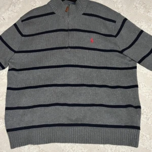 Grå randig tröja från Polo Ralph Lauren - Säljer en stilren grå tröja från Polo Ralph Lauren med mörkblå ränder och en halv dragkedja framtill. Tröjan har långa ärmar och ribbade detaljer vid krage, ärmslut och nederkant. Perfekt för en avslappnad och snygg look.