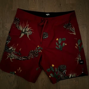 Röda shorts med mönster från Vans - Snygga röda shorts från Vans med coolt mönster av kaktusar och ormar. De har en praktisk snörning i midjan och är perfekta för sommarens äventyr. Shortsen har en normal passform och är tillverkade i ett bekvämt material. Badshorts!