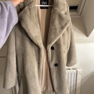 Beige pälsjacka från Zara - Säljer en snygg beige pälsjacka från Zara. Jackan har en klassisk design med stora knappar framtill och en mjuk, fluffig yta. Perfekt för att hålla sig varm och stilren under kyliga dagar.