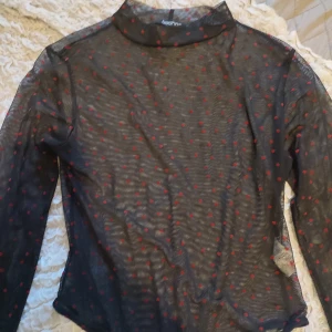 Svart mesh topp med röda prickar från Boohoo - Snygg svart mesh topp från Boohoo med röda prickar. Toppen har långa ärmar och en hög krage, perfekt för en trendig look. Materialet är lätt och genomskinligt vilket ger en cool stil.