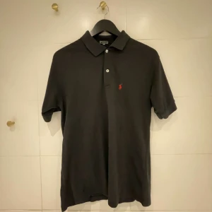 Svart pikétröja från Polo Ralph Lauren - Snygg svart pikétröja från Polo Ralph Lauren med klassisk krage och knappslå. Den har en liten röd broderad logotyp på bröstet. Perfekt för en stilren look.