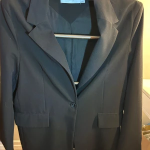 Svart kavaj - Elegant svart kavaj med klassisk design. Kavajen har en enkel knapp framtill och två fickor. Perfekt för en stilren look. Har axel vadd och en slits baktill. 