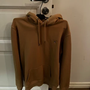Beige hoodie från GANT - Säljer en snygg beige hoodie från GANT med broderad logga på bröstet. Den har en klassisk känguruficka och justerbar huva med snören. Perfekt för en avslappnad stil. 🧥