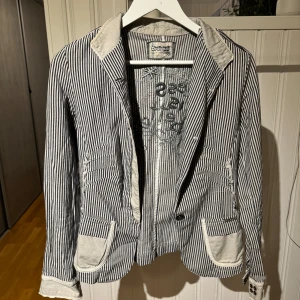 Randig kavaj från Desigual - Säljer en snygg randig kavaj från Desigual i blått och vitt. Kavajen har en unik design med broderade detaljer på insidan och kontrasterande fickor och krage. Perfekt för dig som vill sticka ut med stil!