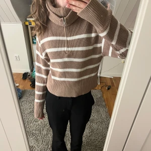 Beige och vit randig stickad tröja - Mysig stickad tröja i beige och vitt med randigt mönster. Tröjan har en hög krage med dragkedja och långa ärmar. Perfekt för kyligare dagar.