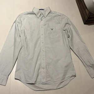 Vit prickig skjorta från GANT - Snygg vit skjorta från GANT med små svarta och blåa prickar och klassisk krage. Skjortan har en bröstficka med broderad logotyp och knappar framtill. Perfekt för en stilren look. Storleken är M. Pris kan diskuteras vid snabb affär! 