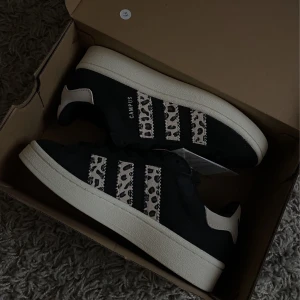 Svarta Adidas Campus med leopardmönster - Snygga svarta Adidas Campus sneakers med leopardmönstrade detaljer på de klassiska tre ränderna. Skorna har en vit sula och svart snörning, vilket ger en cool kontrast. Perfekta för dig som vill ha en unik twist på en klassisk modell. originalpris 250€