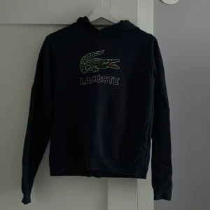 Svart hoodie från Lacoste - Snygg svart hoodie från Lacoste med det ikoniska krokodilmärket på bröstet. Perfekt för en avslappnad stil. Den har långa ärmar och en klassisk huva. 