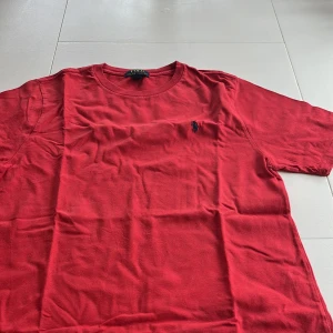 Röd t-shirt från Ralph Lauren - Säljer en klassisk röd t-shirt från Ralph Lauren med broderad logga på bröstet. T-shirten är kortärmad och tillverkad i bomull, perfekt för en avslappnad stil.