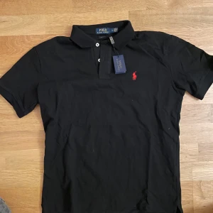 Svart pikétröja från Ralph Lauren - Säljer en klassisk svart pikétröja från Ralph Lauren med den ikoniska röda loggan på bröstet. Tröjan har en krage med knappar och korta ärmar. Perfekt för en stilren look.