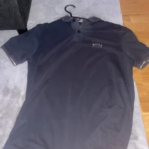 Svart kortärmad t-shirt från Hugo Boss - Säljer en stilren svart t-shirt från Hugo Boss i slim fit. T-shirten har korta ärmar och en klassisk krage med knappar. Perfekt för en snygg och avslappnad look.