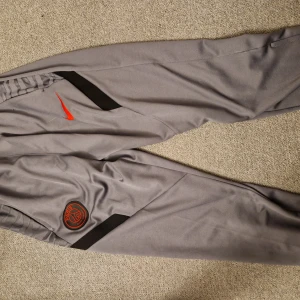 Grå träningsbyxor från Nike psg - Snygga grå träningsbyxor från Nike med svart och röd detalj. Byxorna har en elastisk midja och en röd Nike-logga på sidan. Perfekta för sport och fritid.