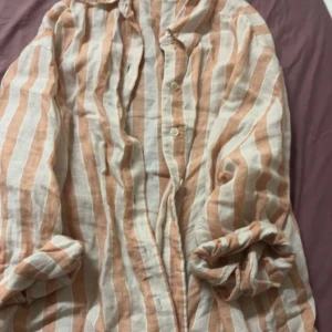 Randig linneskjorta och shorts från H&M - Säljer ett set med randig skjorta och shorts i linne från H&M. Skjortan har långa ärmar och knappar framtill. Färgerna är beige och vit, perfekt för en avslappnad stil.