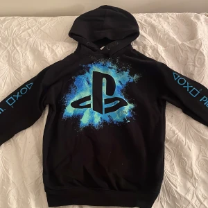 Svart PlayStation hoodie från H&M - Cool svart hoodie från H&M med PlayStation-logga i blått och vitt på framsidan. Ärmarna har PlayStation-symboler i blått. Perfekt för gaming eller chill dagar. 🕹️