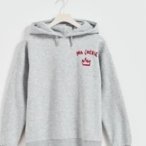 Grå hoodie med röd text - Säljer en grå hoodie med texten 'MA CHÉRIE' och en krona i rött på bröstet och ryggen. Den har en klassisk huva med dragsko och långa ärmar. Perfekt för en avslappnad stil.
